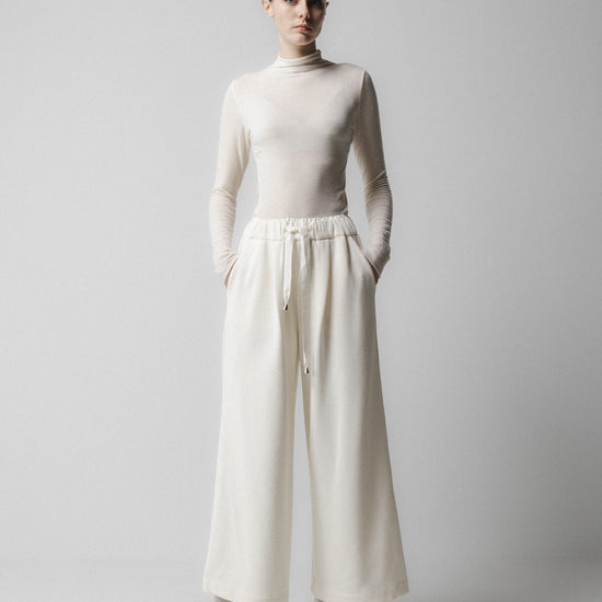 Person wearing a white turtleneck and wide-leg pants on a plain  background Emin + Paul
