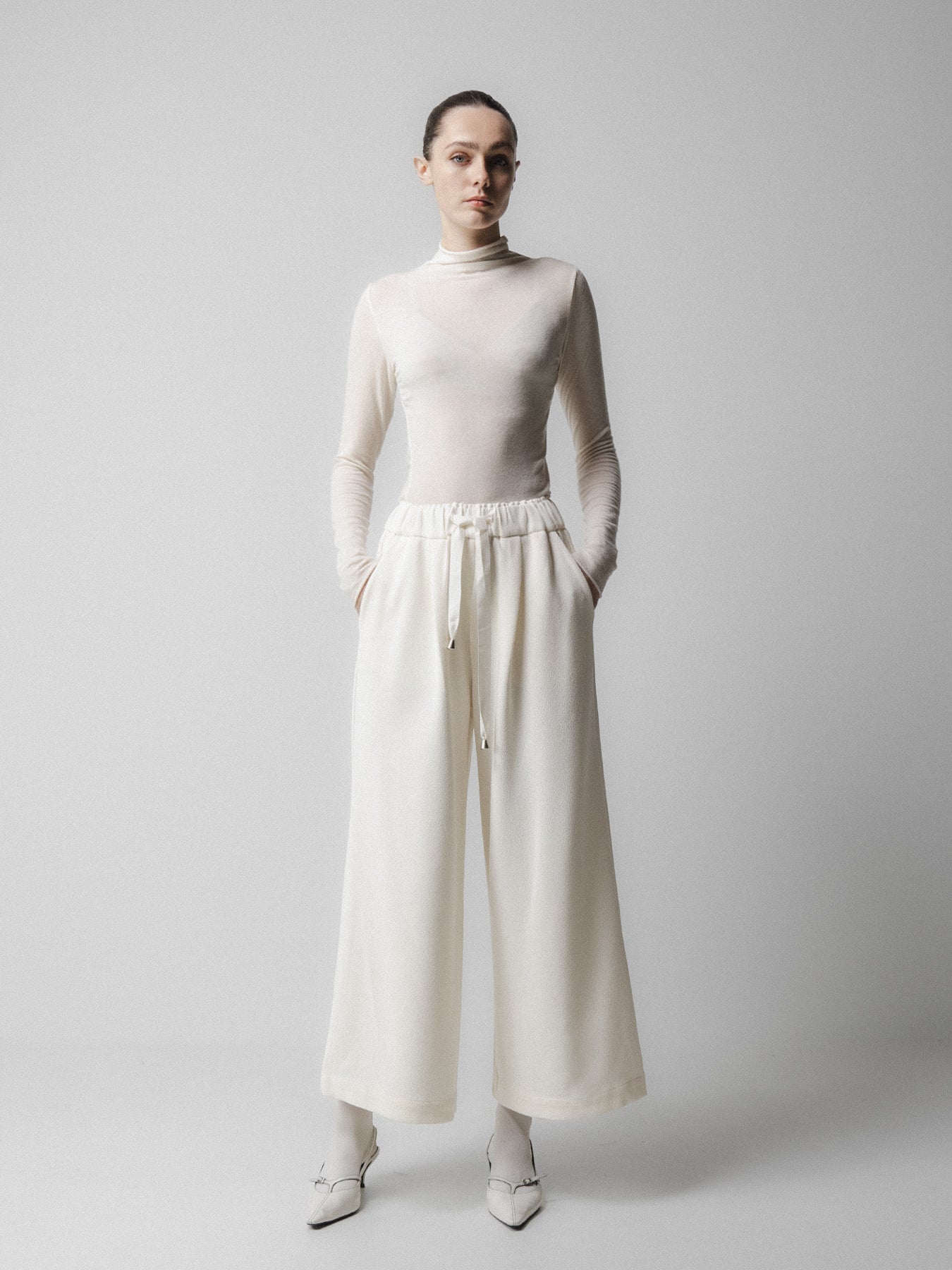 Person wearing a white turtleneck and wide-leg pants on a plain  background Emin + Paul