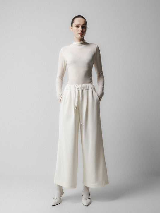Person wearing a white turtleneck and wide-leg pants on a plain  background Emin + Paul