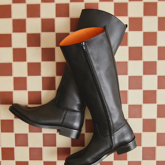 A pair of black leather knee-high riding boots with side zippers, displayed on a red and cream checkered tile floor. The interior lining of the boots is a vibrant orange.