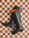 A pair of black leather knee-high riding boots with side zippers, displayed on a red and cream checkered tile floor. The interior lining of the boots is a vibrant orange.