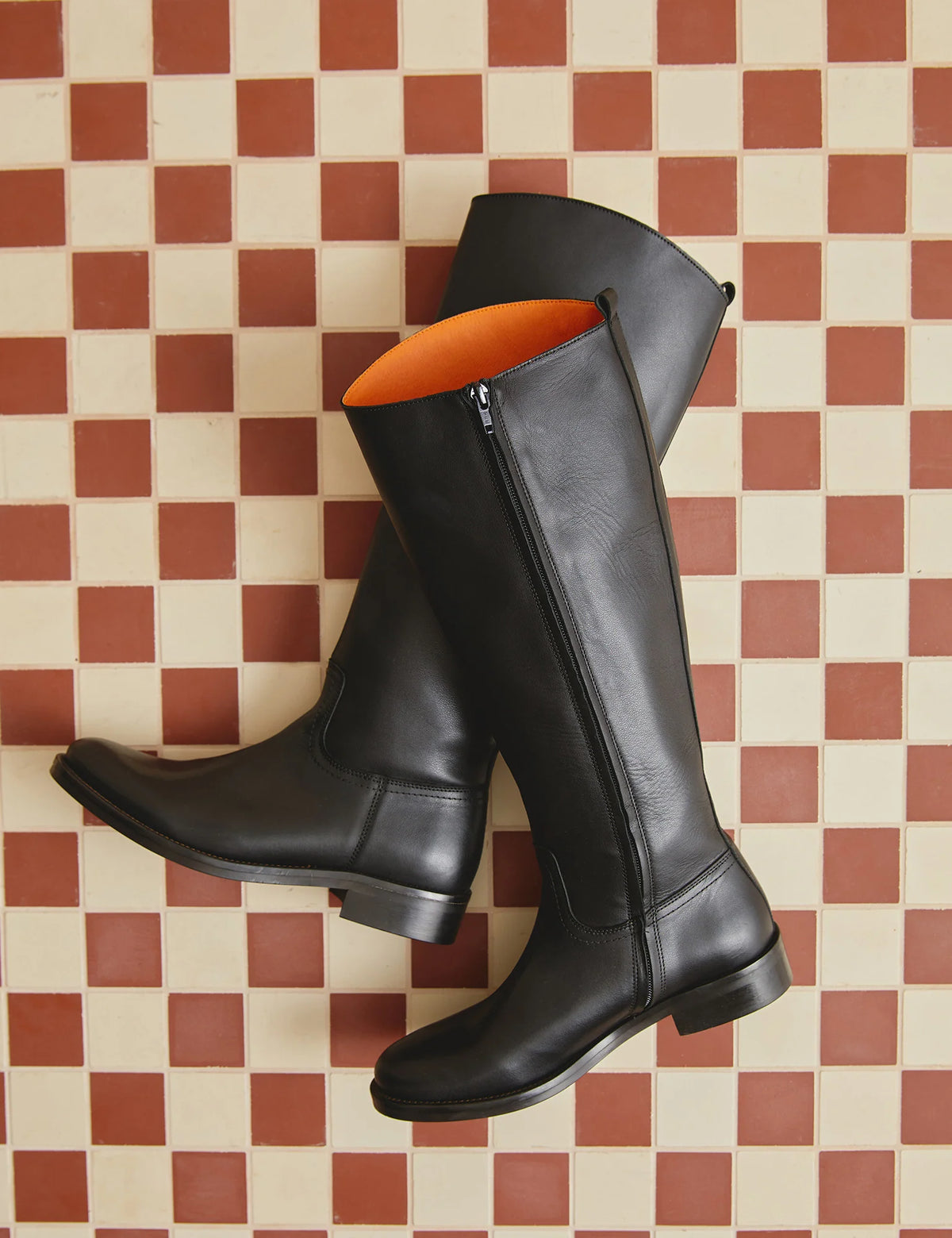 A pair of black leather knee-high riding boots with side zippers, displayed on a red and cream checkered tile floor. The interior lining of the boots is a vibrant orange.