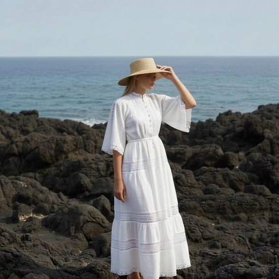 Woman in a white dress and straw hat standing on rocky terrain by the ocean.