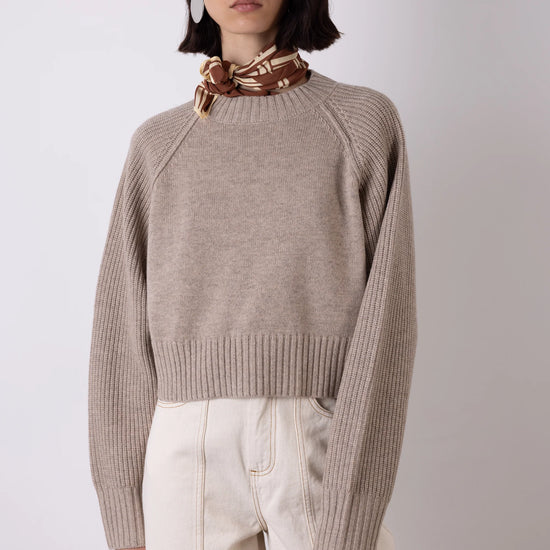 Woman wearing a beige sweater and white pants with a brown scarf on a plain background