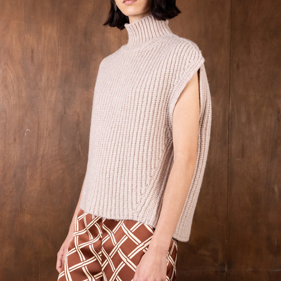 Woman wearing a beige ribbed turtleneck sleeveless sweater against a wooden background