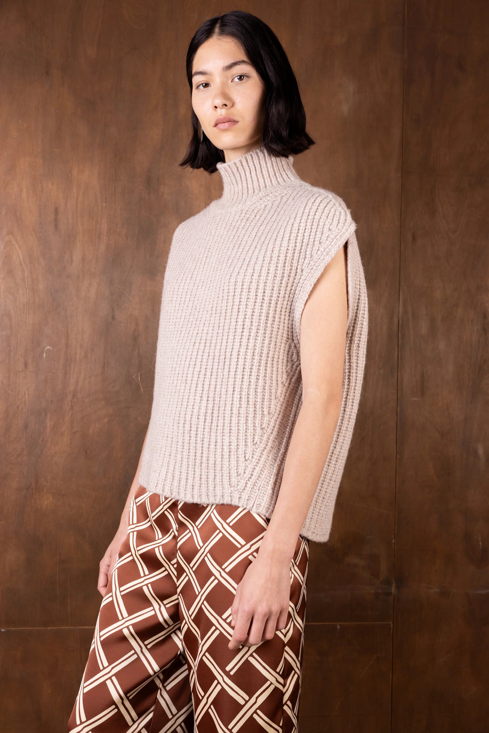 Woman wearing a beige ribbed turtleneck sleeveless sweater against a wooden background