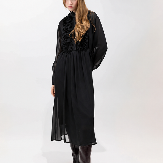 A woman modeling a long black midi dress with a high ruffled collar and sheer long sleeves, paired with dark brown pointed-toe boots, against a plain white studio background.
