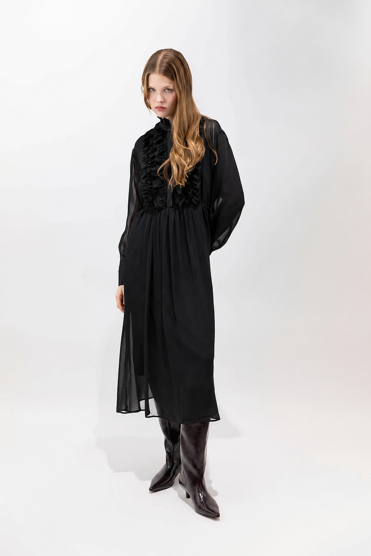 A woman modeling a long black midi dress with a high ruffled collar and sheer long sleeves, paired with dark brown pointed-toe boots, against a plain white studio background.