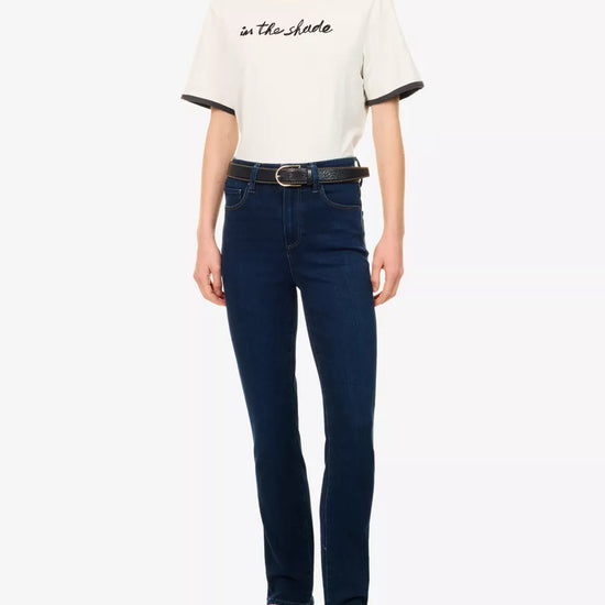 A model wearing a white short-sleeve t-shirt with black lettering on the chest and black ringer trim on the sleeves, paired with dark blue skinny jeans, a black belt, and dark brown leather flats, against a plain white background.