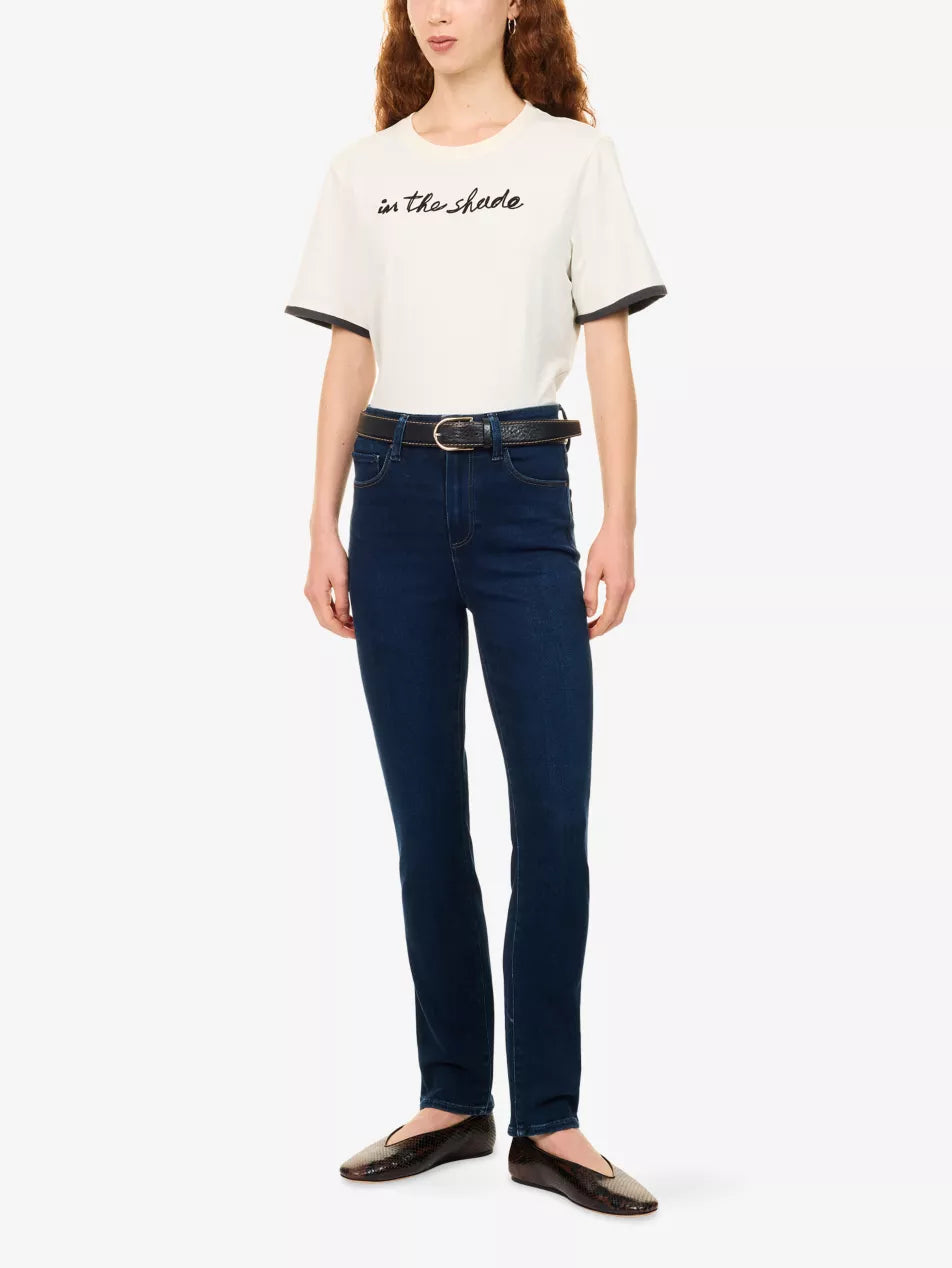 A model wearing a white short-sleeve t-shirt with black lettering on the chest and black ringer trim on the sleeves, paired with dark blue skinny jeans, a black belt, and dark brown leather flats, against a plain white background.