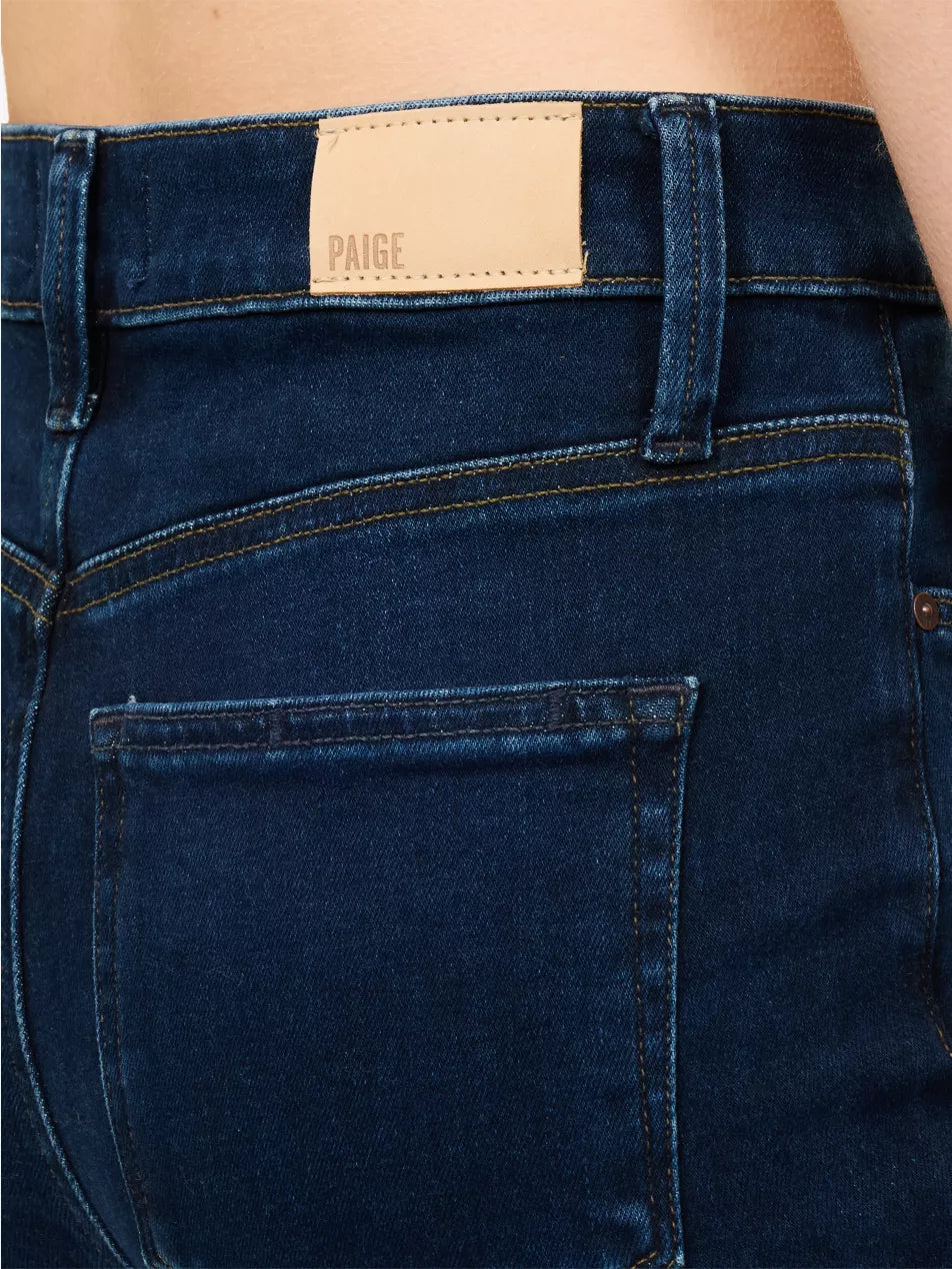 A model wearing a white short-sleeve t-shirt with black lettering on the chest and black ringer trim on the sleeves, paired with dark blue skinny jeans, a black belt, and dark brown leather flats, against a plain white background.