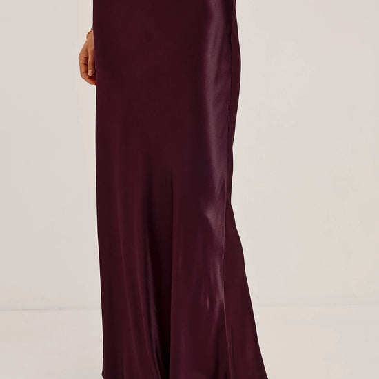 A model wearing a floor-length, satin-like dark plum skirt with a draped, fluid silhouette and black flat sandals, shown from the waist down against a plain light gray studio background.