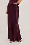 A model wearing a floor-length, satin-like dark plum skirt with a draped, fluid silhouette and black flat sandals, shown from the waist down against a plain light gray studio background.
