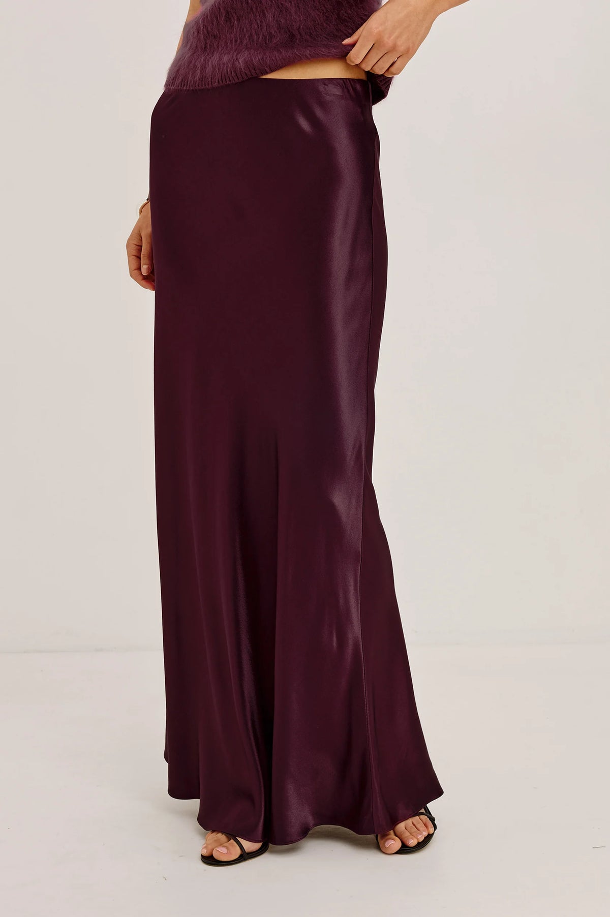 A model wearing a floor-length, satin-like dark plum skirt with a draped, fluid silhouette and black flat sandals, shown from the waist down against a plain light gray studio background.