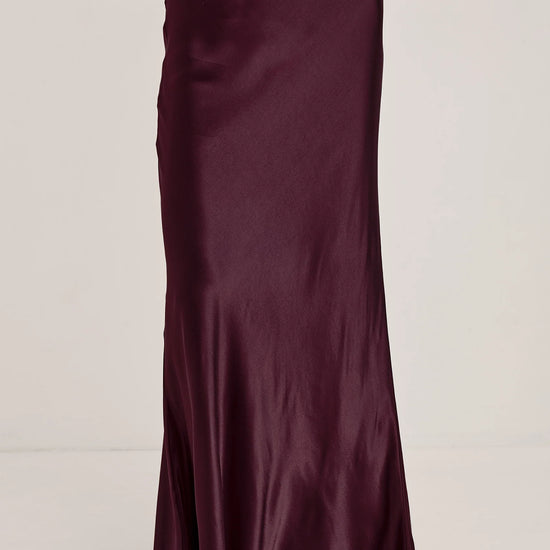 A woman wearing a long, floor-length satin maxi skirt in deep burgundy. The skirt has a subtle sheen and is styled with a matching knit top and black flat sandals against a plain light gray studio background.