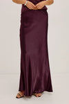 A woman wearing a long, floor-length satin maxi skirt in deep burgundy. The skirt has a subtle sheen and is styled with a matching knit top and black flat sandals against a plain light gray studio background.