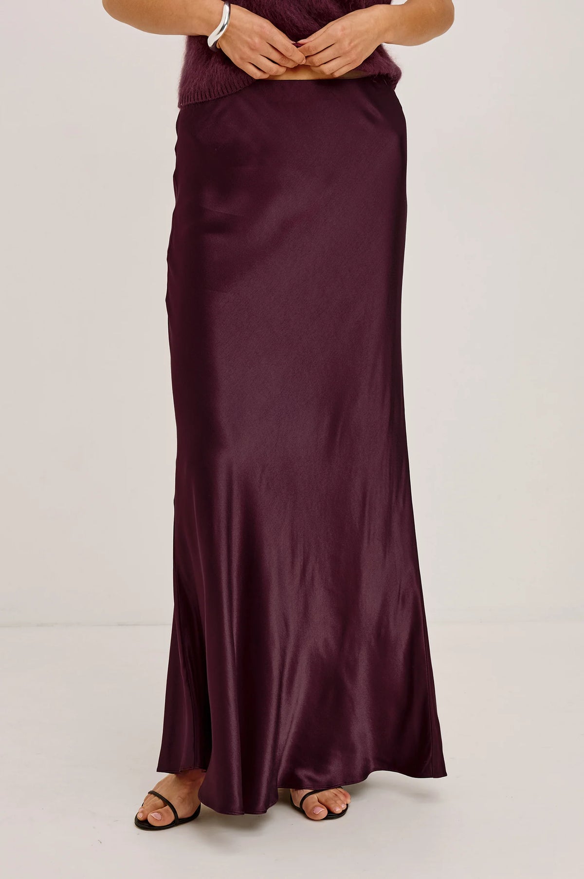 A woman wearing a long, floor-length satin maxi skirt in deep burgundy. The skirt has a subtle sheen and is styled with a matching knit top and black flat sandals against a plain light gray studio background.
