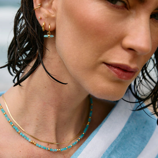 Woman wearing gold earrings and a turquoise beaded necklace with a blurred background