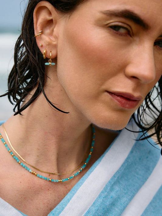 Woman wearing gold earrings and a turquoise beaded necklace with a blurred background
