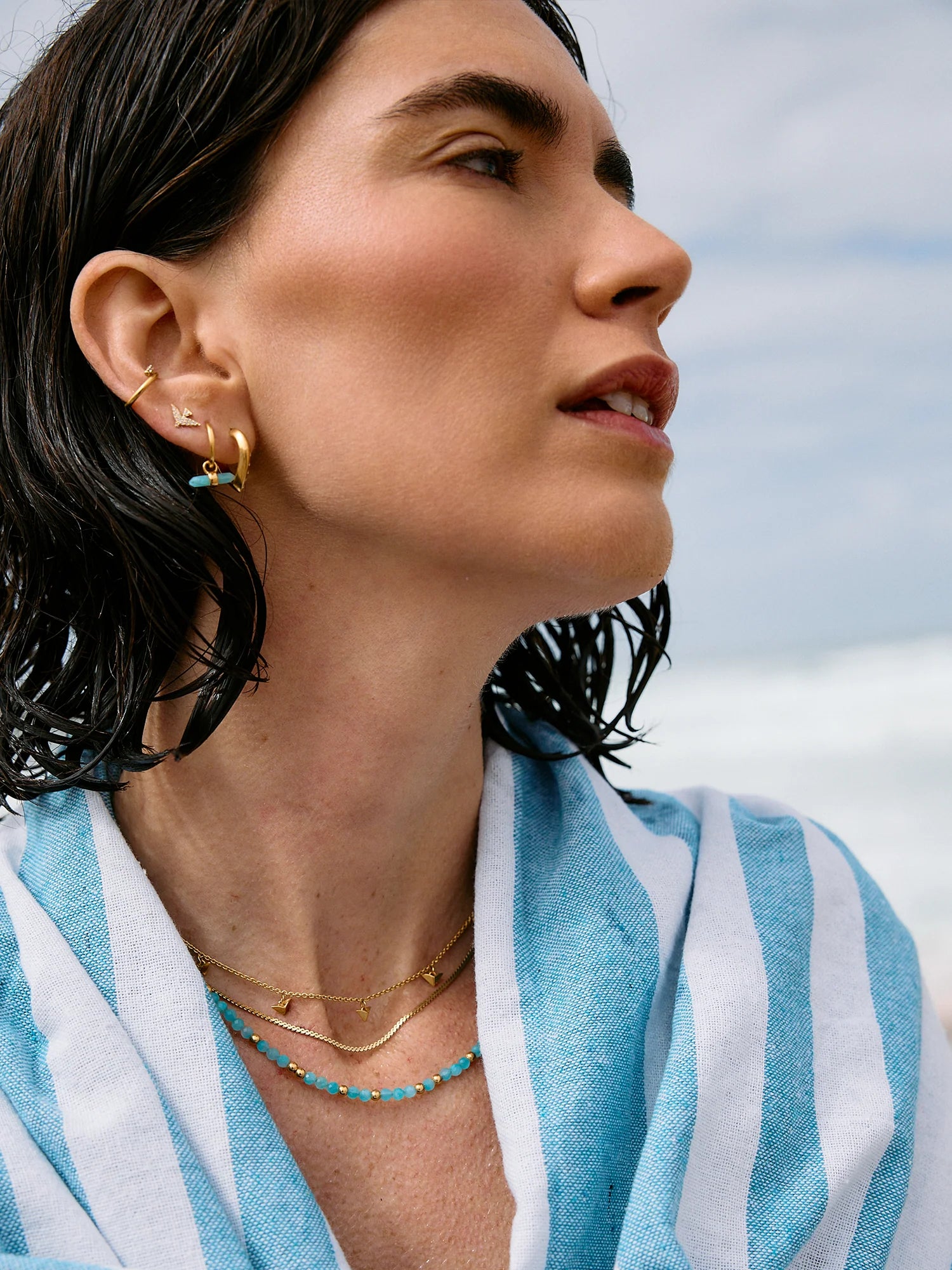 Woman wearing gold earrings and necklaces with a blue and white striped garment against a sky background