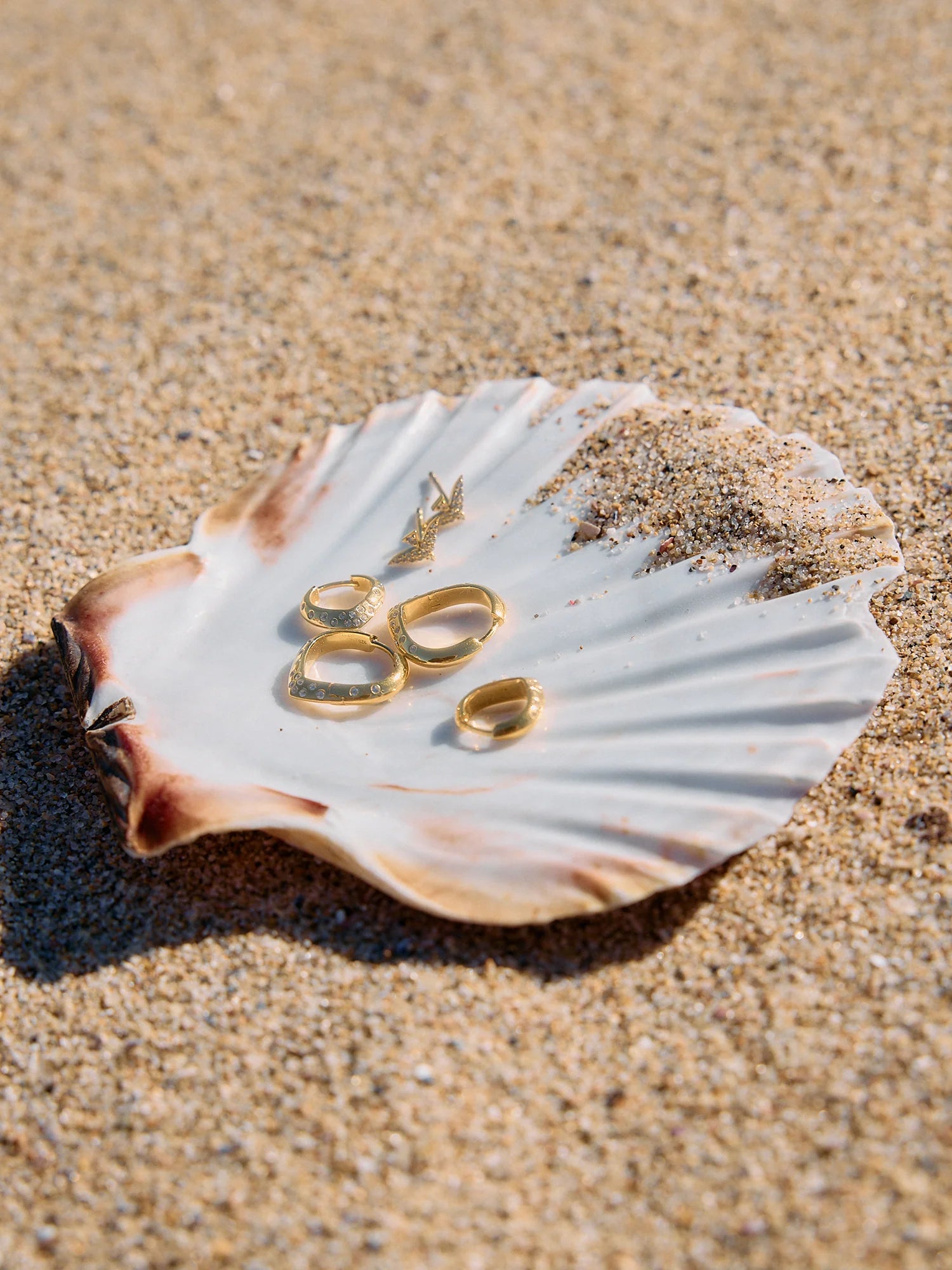 Gold rings and rachel jackson earrings on a seashell with sand in the background