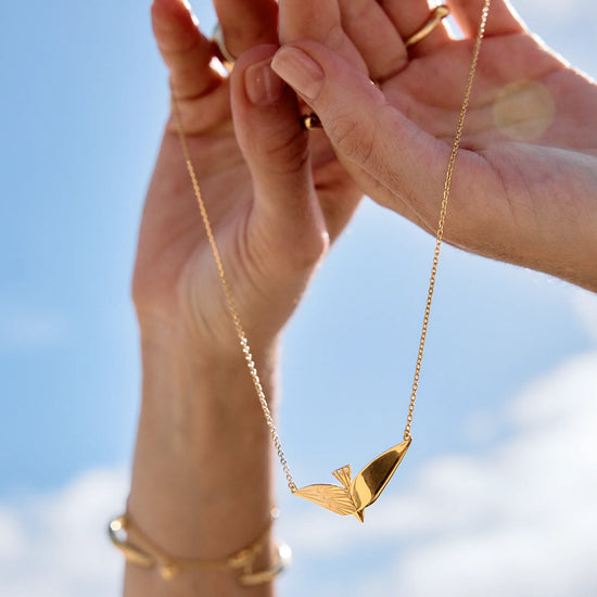 Gold necklace with a leaf pendant held by hands against a blue sky.