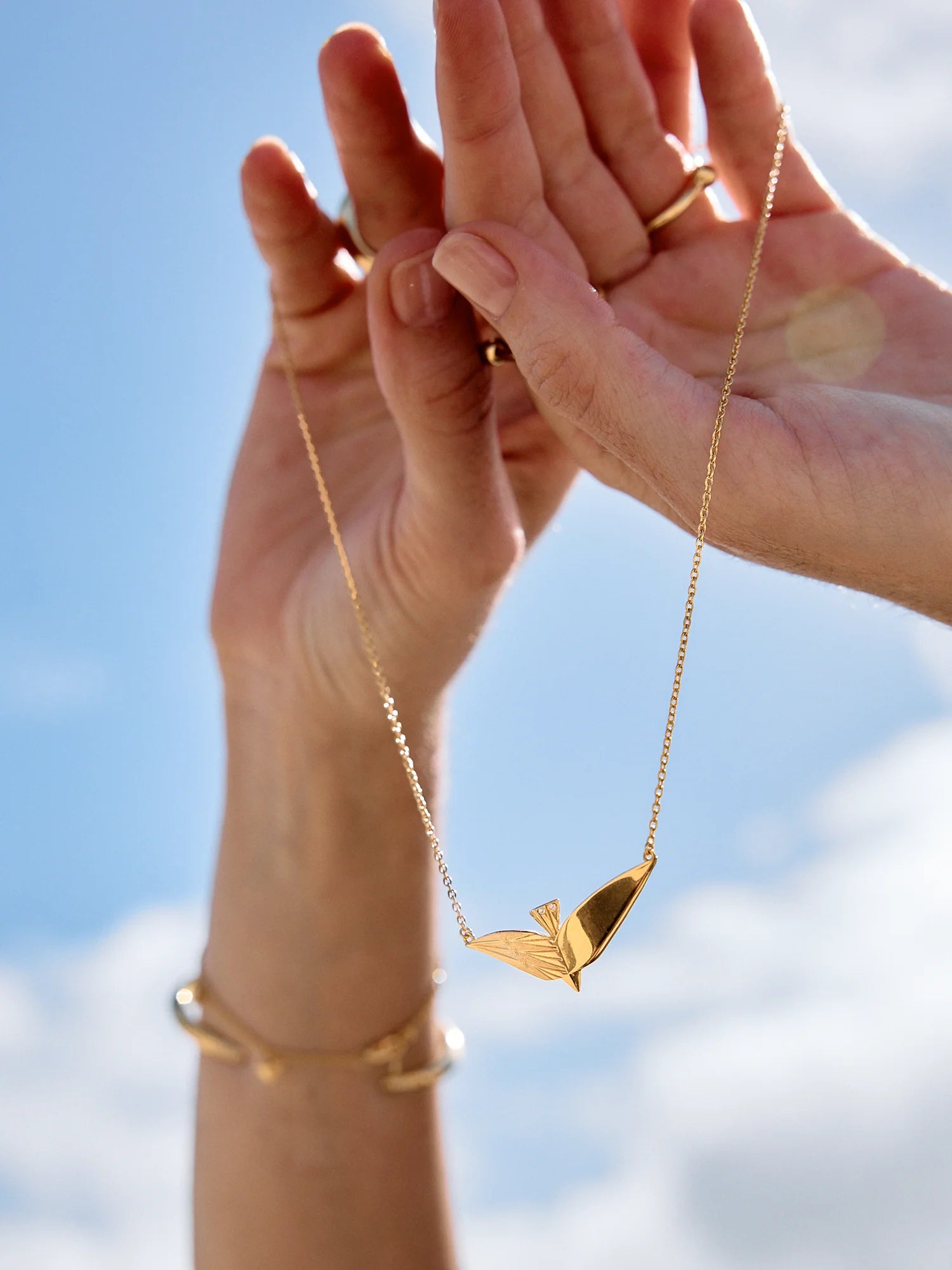 Gold necklace with a leaf pendant held by hands against a blue sky.