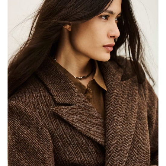 A woman wearing a brown herringbone patterned coat over a brown collared shirt, photographed against a plain light background.