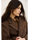 A woman wearing a brown herringbone patterned coat over a brown collared shirt, photographed against a plain light background.