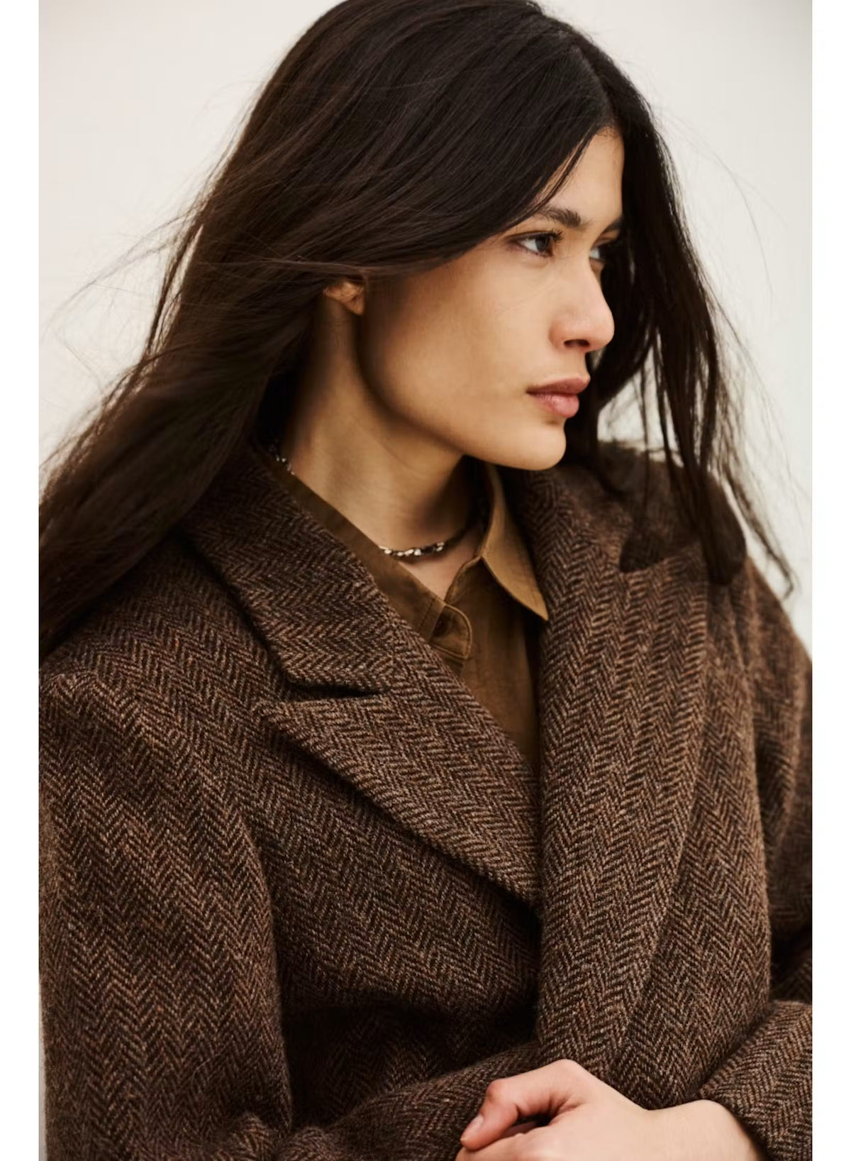 A woman wearing a brown herringbone patterned coat over a brown collared shirt, photographed against a plain light background.