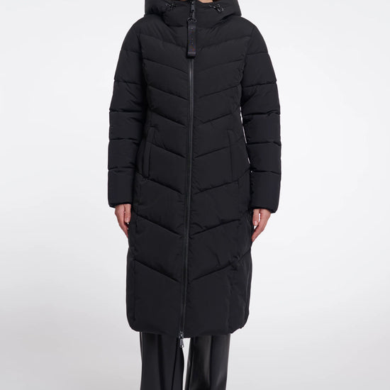 A woman modeling a long black hooded puffer coat with a front zipper, paired with black pants, against a plain light gray studio background.