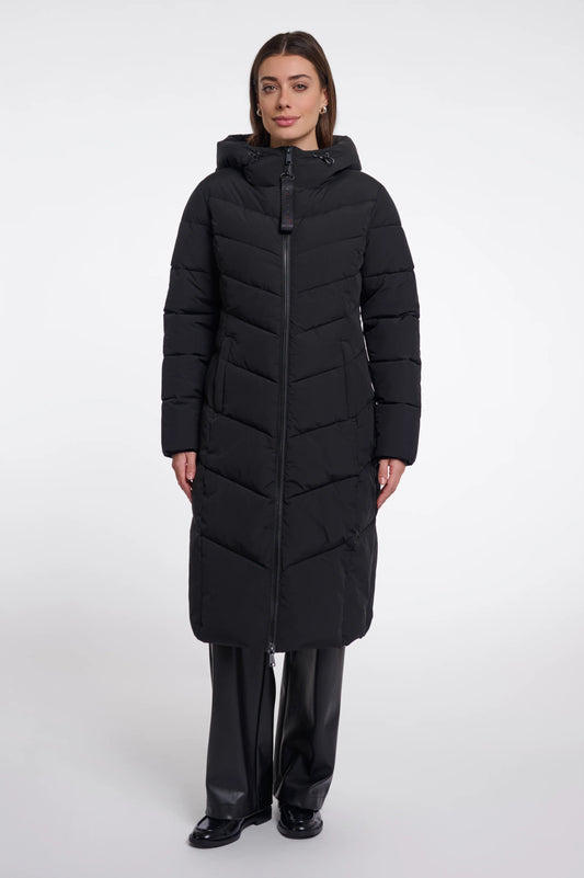 A woman modeling a long black hooded puffer coat with a front zipper, paired with black pants, against a plain light gray studio background.