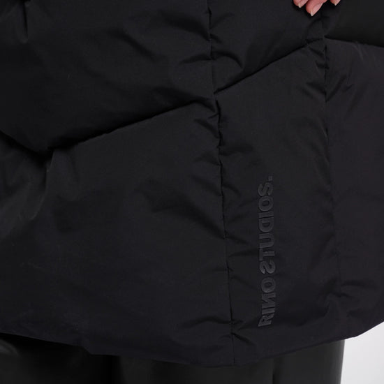 Close-up of a black puffer jacket with horizontal quilting and a vertical tonal 'SODIUTS AMIR' logo on the lower right side, worn with black pants. A hand is visible at the top right.