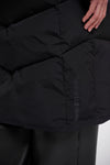 Close-up of a black puffer jacket with horizontal quilting and a vertical tonal 'SODIUTS AMIR' logo on the lower right side, worn with black pants. A hand is visible at the top right.