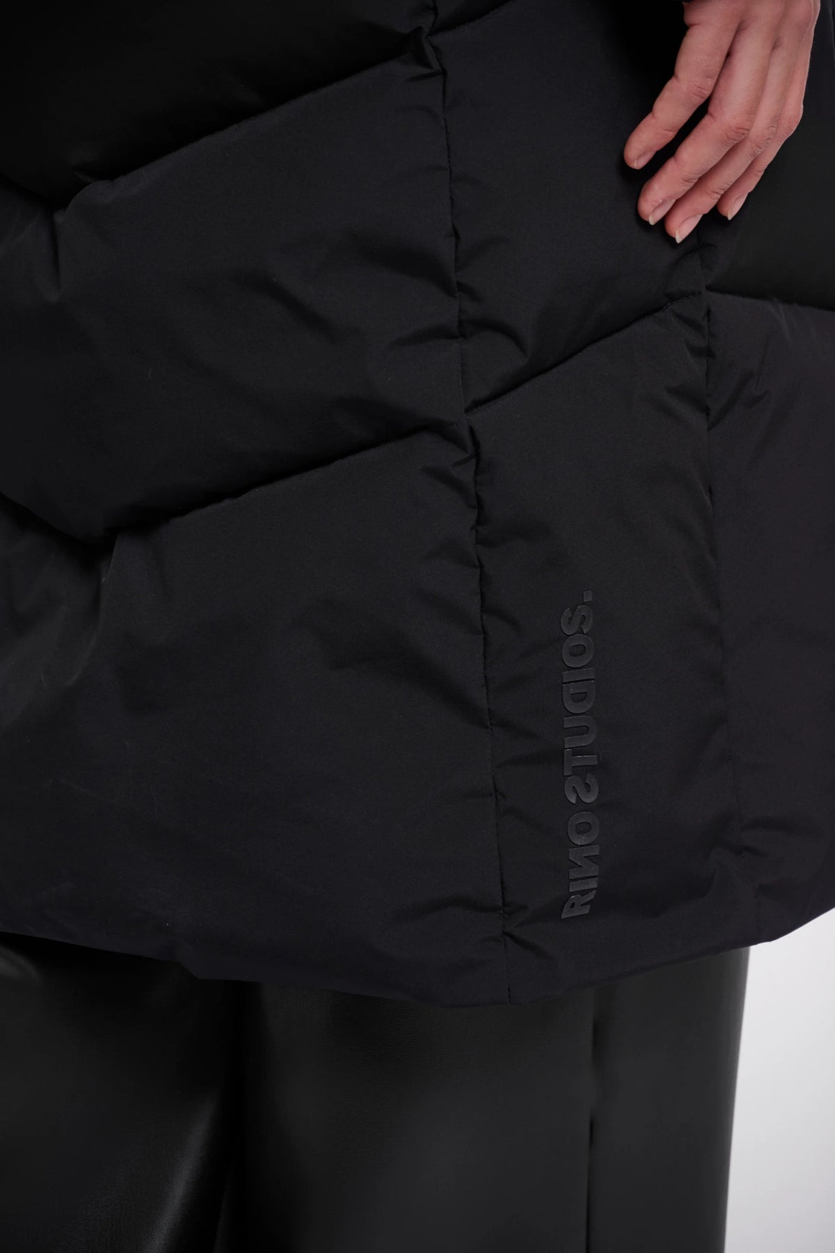 Close-up of a black puffer jacket with horizontal quilting and a vertical tonal 'SODIUTS AMIR' logo on the lower right side, worn with black pants. A hand is visible at the top right.