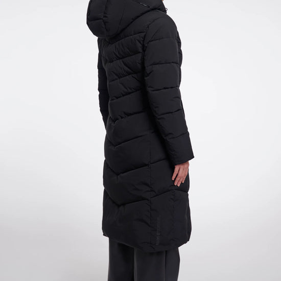 A model shown from the back wearing a long black puffer coat with a hood, paired with dark wide-leg trousers, against a plain light gray studio background.
