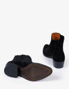 A pair of black suede ankle boots shown from the side and back on a clean white background, displaying the black sole and a contrasting orange-brown interior lining.