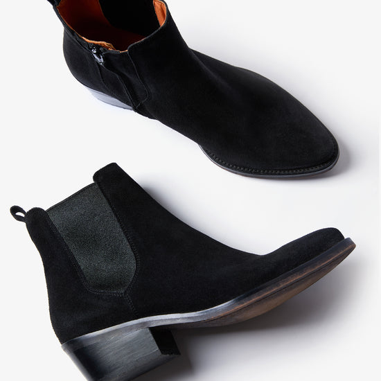 A pair of black suede Chelsea-style ankle boots with elastic side panels and low block heels, shown from side and angled top views on a light gray background.