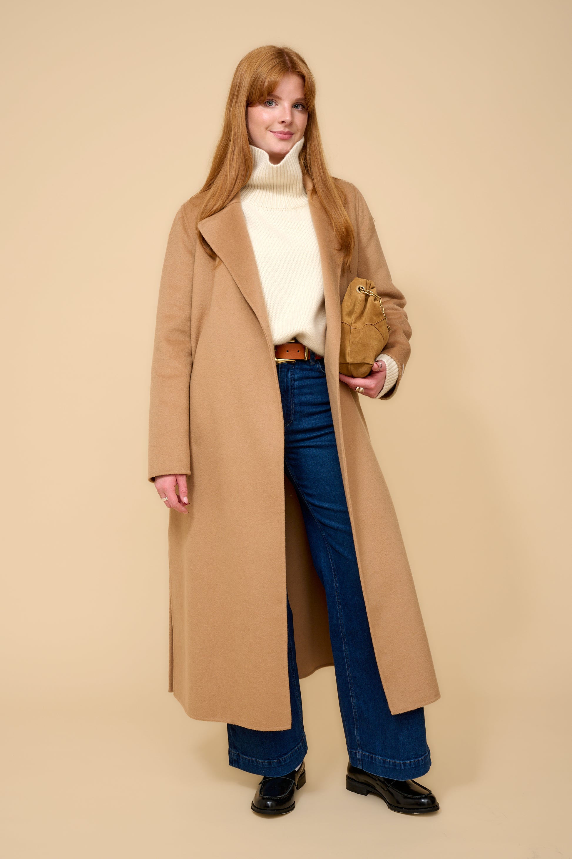 model shot wearing camel edge to edge wool coat, cream knit, blue jeans holding a camel handbag