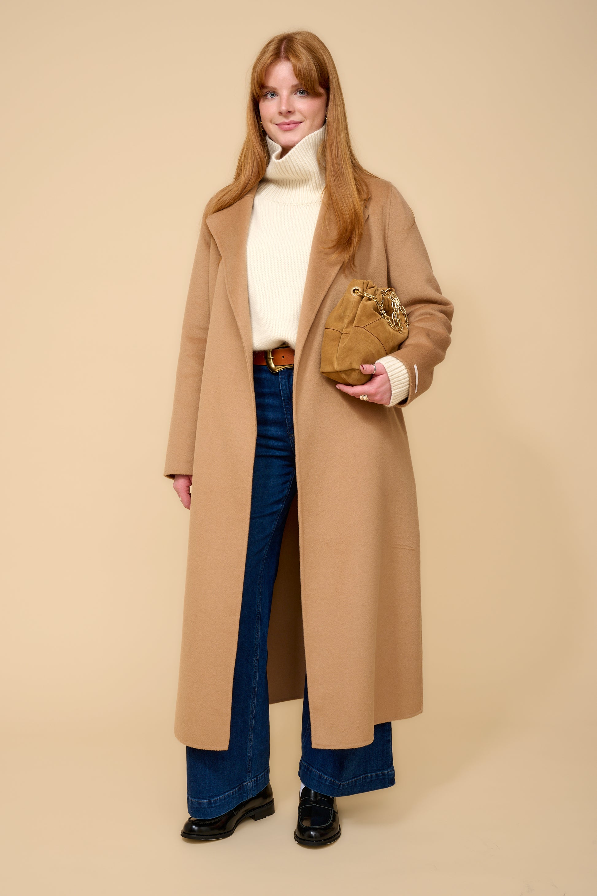 model shot wearing cream thick roll neck knit, denim jeans, camel coat and brown belt.