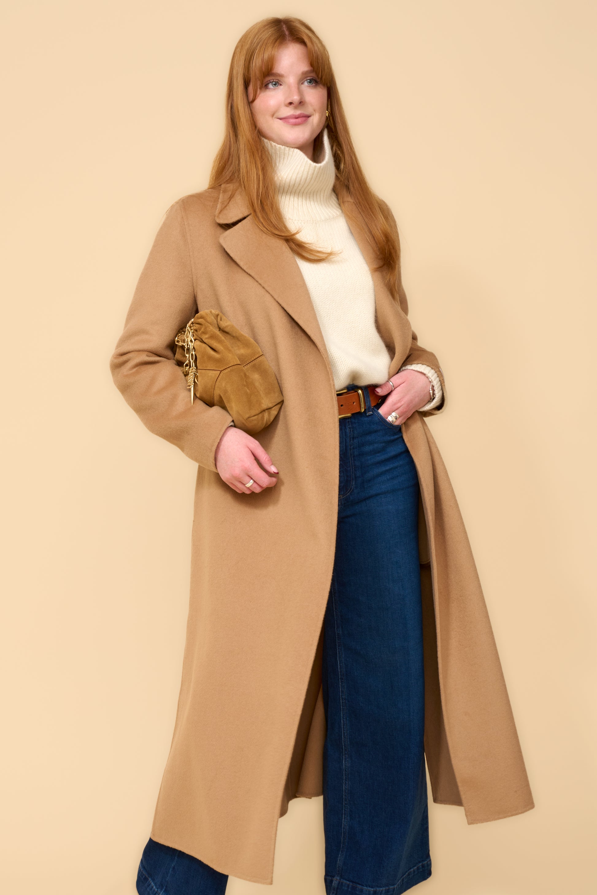model shot wearing camel edge to edge wool coat, cream knit, blue jeans holding a camel handbag