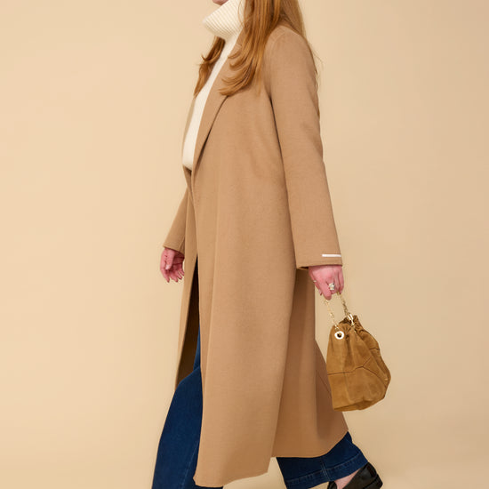 model shot wearing camel edge to edge wool coat, cream knit, blue jeans holding a camel handbag