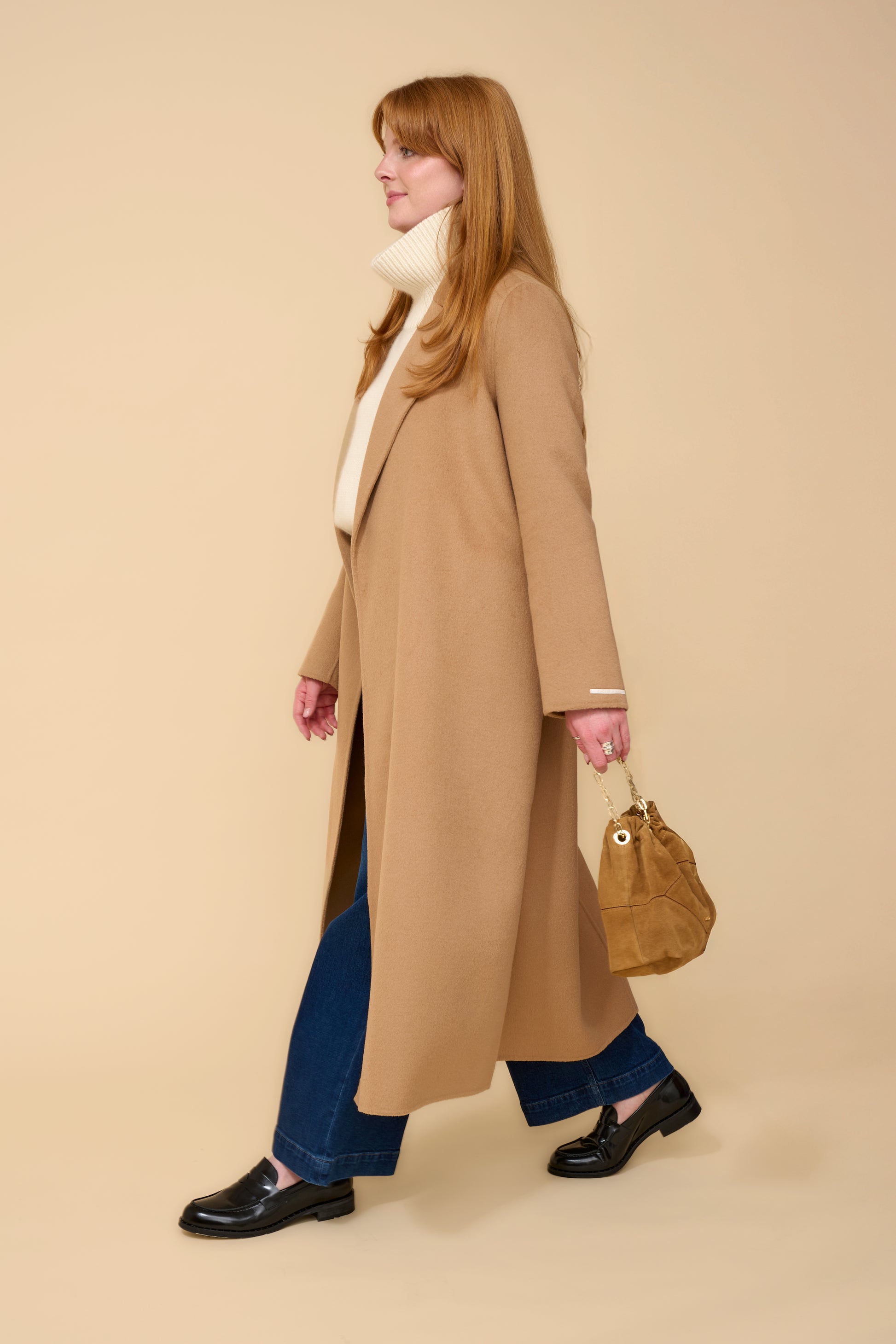 model shot wearing camel edge to edge wool coat, cream knit, blue jeans holding a camel handbag