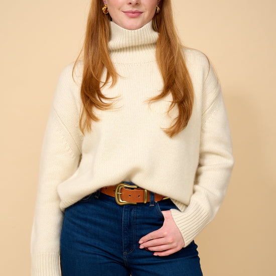 model shot wearing white thick roll neck knit, denim jeans and brown belt.