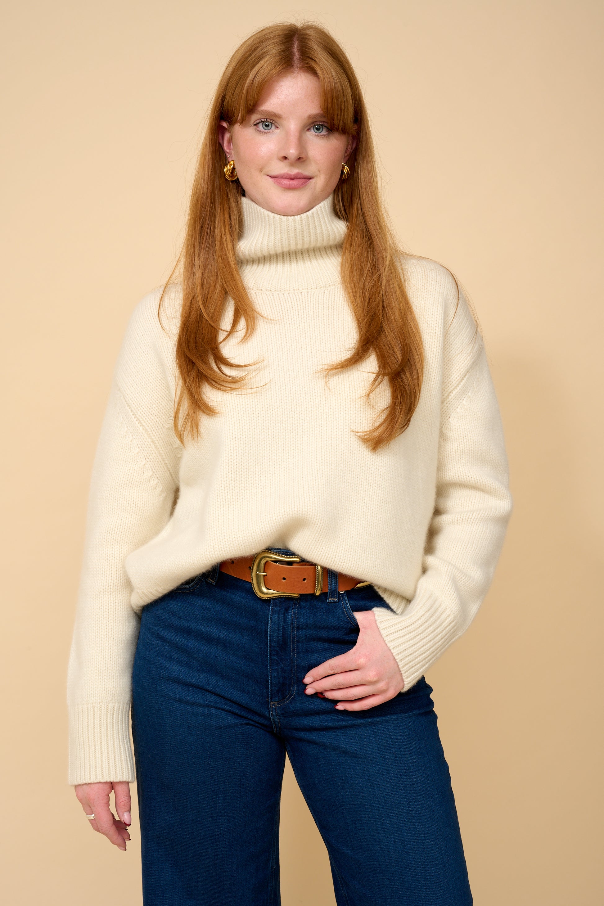 model shot wearing white thick roll neck knit, denim jeans and brown belt.