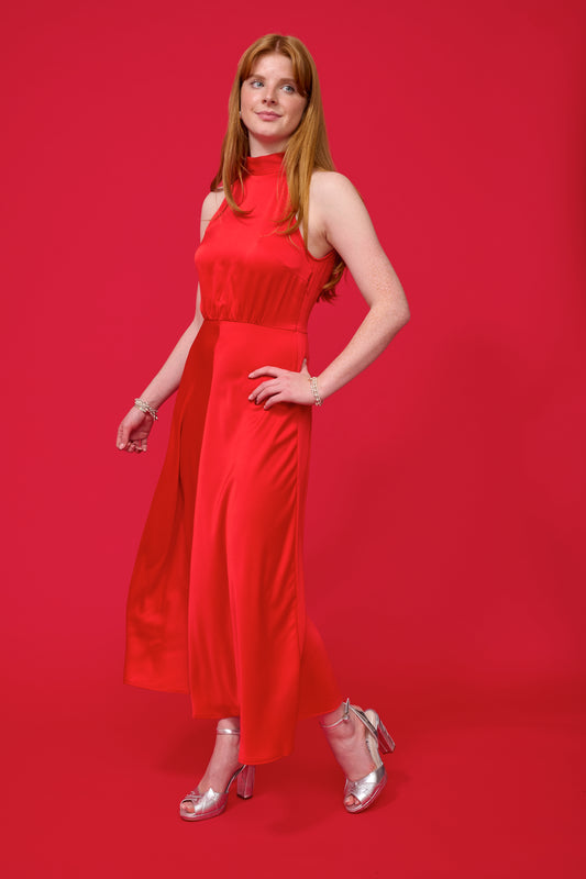 model shot wearing a red halterneck dress with open back and a tie and mid length from samsoe samsoe and silver heels with a red background