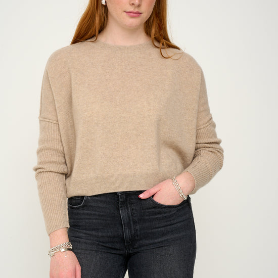 Woman wearing a oatmeal cashmere jumper with round neck and ribbed sleeves, black jeans against a plain background front view