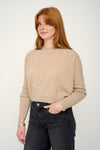 Woman wearing a oatmeal cashmere jumper with round neck and ribbed sleeves, black jeans against a plain background side view
