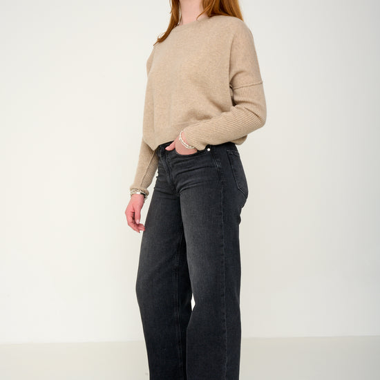 Woman wearing a oatmeal cashmere jumper with round neck and ribbed sleeves, black jeans against a plain background side view