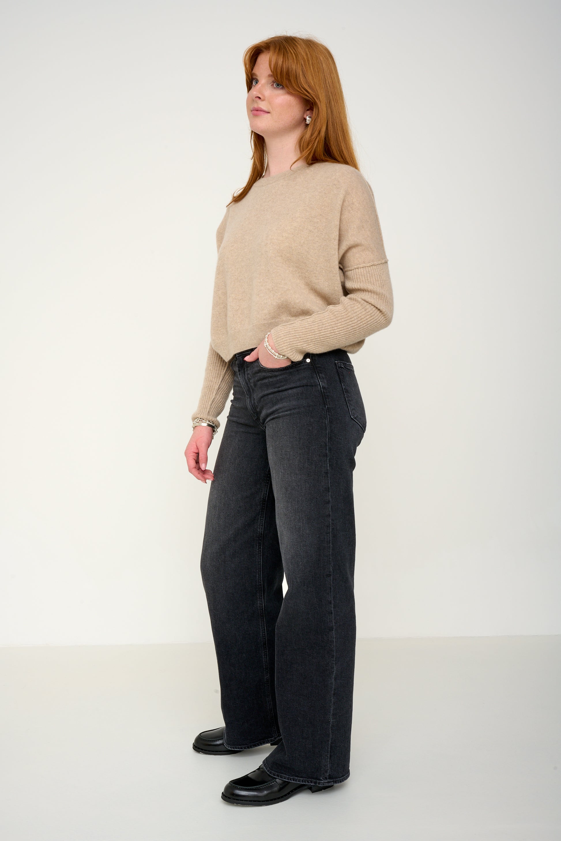 Woman wearing a oatmeal cashmere jumper with round neck and ribbed sleeves, black jeans against a plain background side view